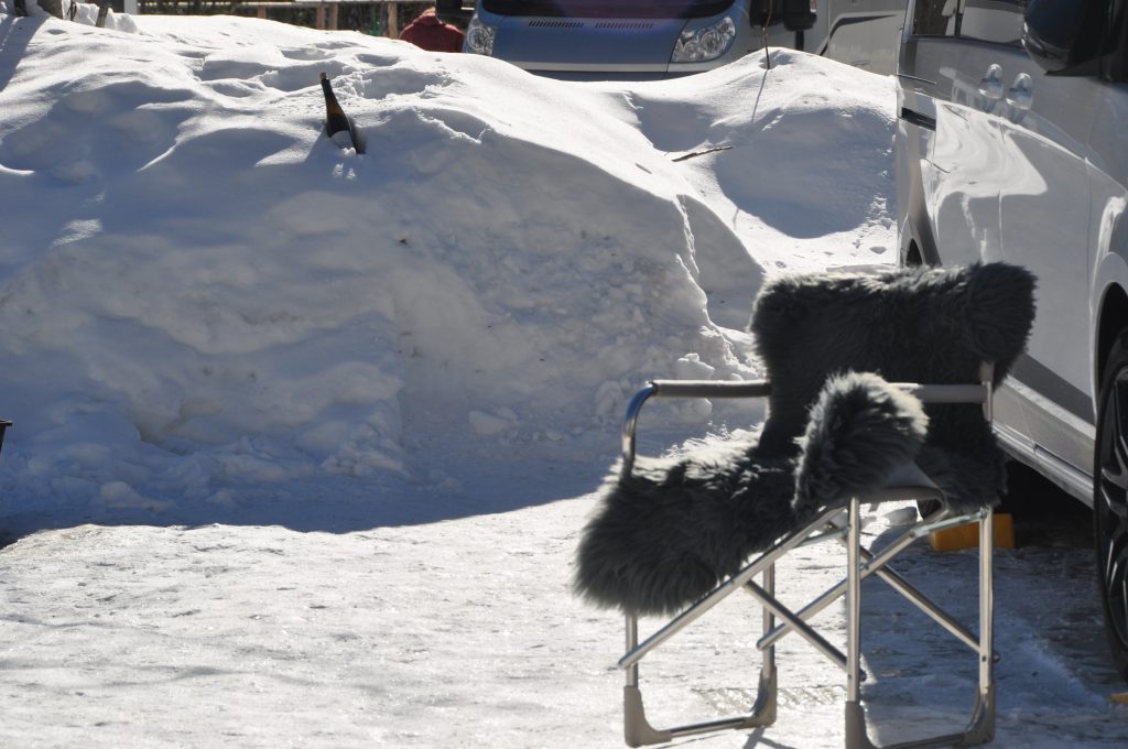Camping-Stuhl mit Fell neben Auto im Schnee. Im Hintergrund steckt eine Flasche Wein im Schnee zum kühlen.